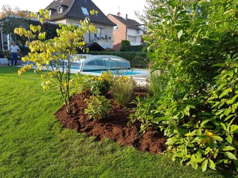 Ein angelegter Garten mit einem gemulchten Blumenbeet, grünem Gras, verschiedenen Sträuchern und einem überdachten Swimmingpool. Im Hintergrund sind Wohnhäuser und zwei Personen zu sehen.