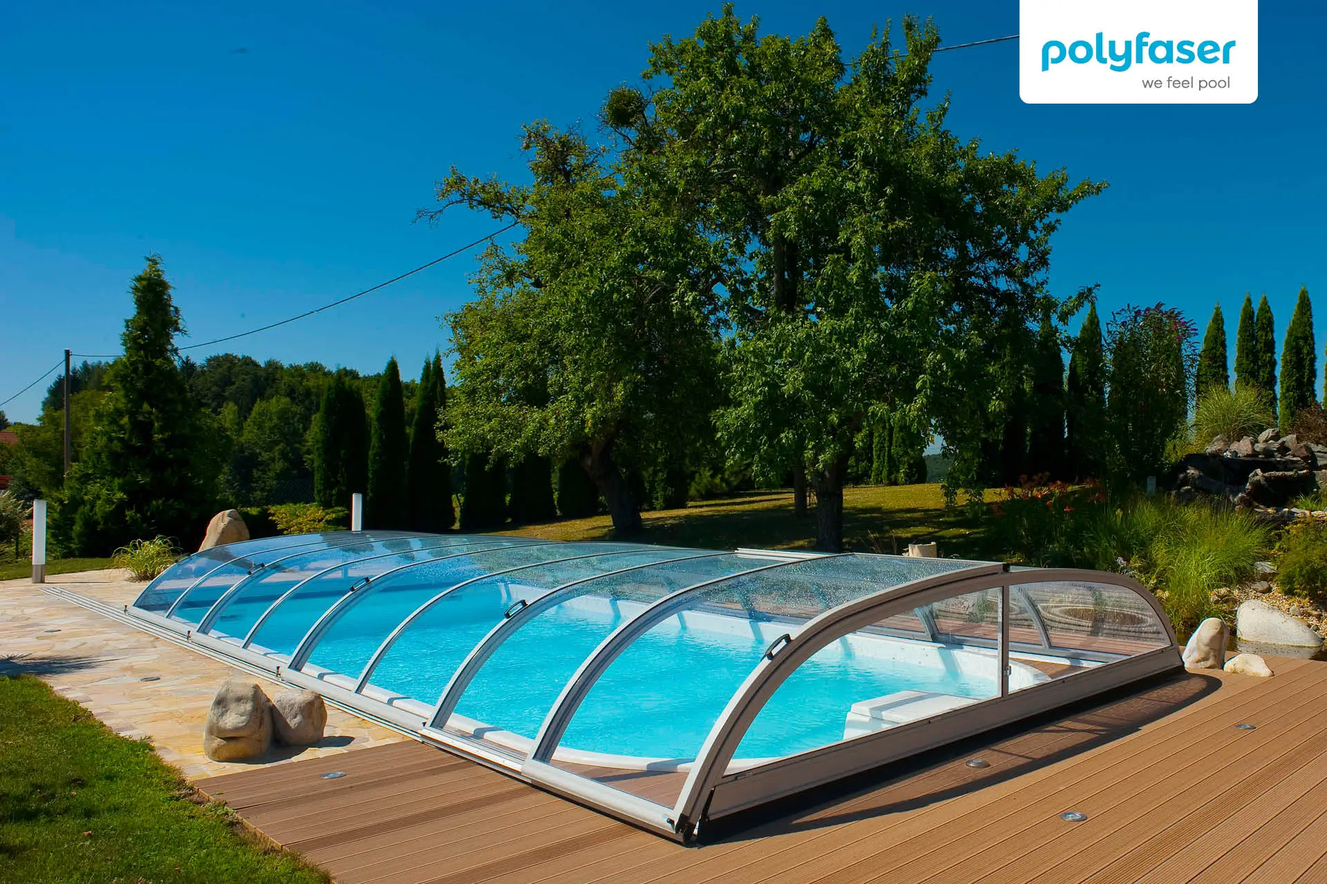 Ein Außenschwimmbecken mit einer transparenten, gewölbten Abdeckung befindet sich auf einer Holzterrasse, umgeben von Gras, Bäumen und Grünanlagen. Der Himmel ist klar und blau. Das Polyfaser-Logo ist in der oberen rechten Ecke zu sehen.