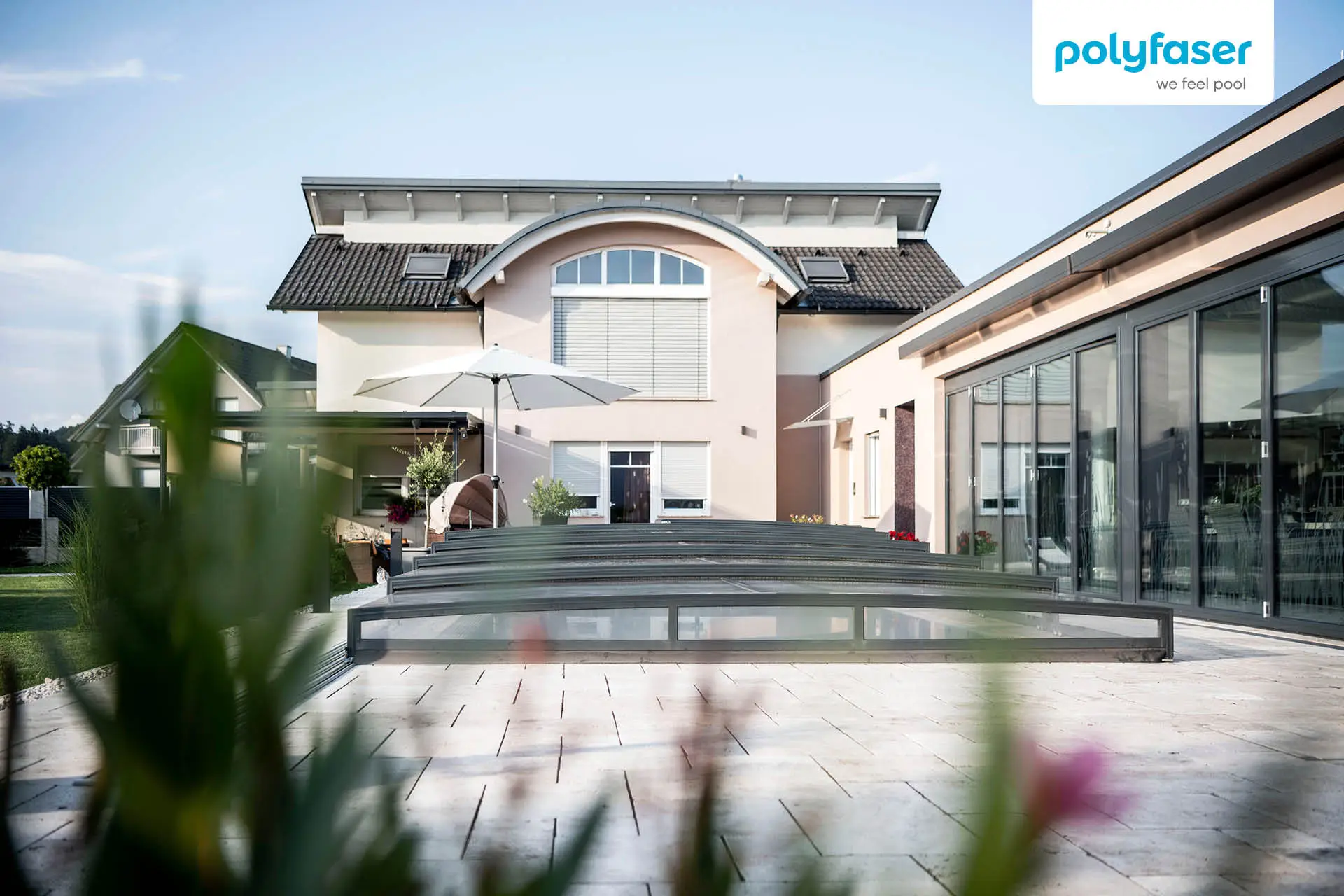 Modernes Haus mit rosafarbenem Äußeren, großen Fenstern und einem überdachten Pool auf einer Steinterrasse. Im Vordergrund sind Grünpflanzen und Blumen zu sehen. Das Polyfaser-Logo ist in der oberen rechten Ecke zu sehen.