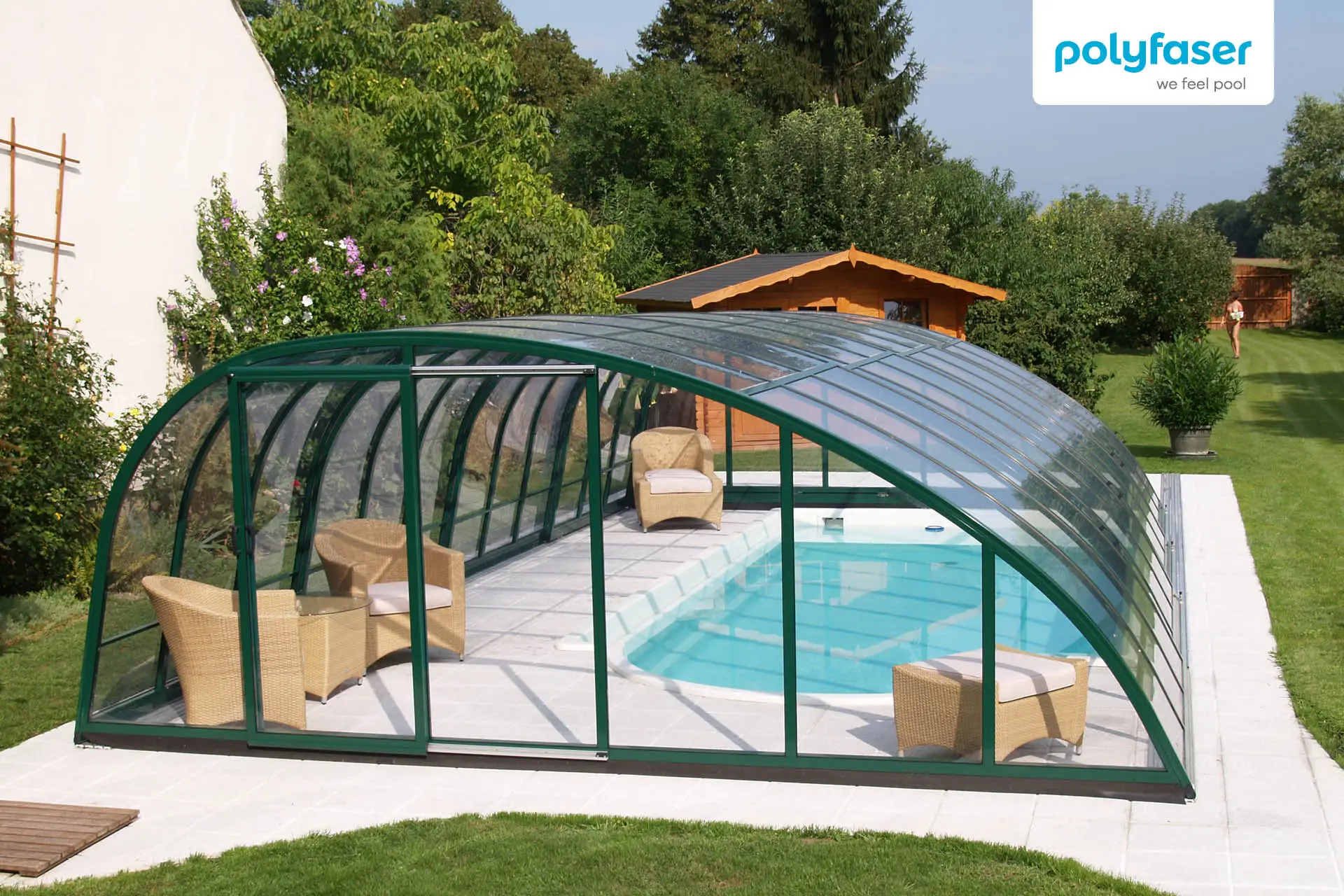 Ein überdachter Außenpool mit einem durchsichtigen Gewölbe, vier Korbstühle mit Kissen neben dem Pool, ein Holzschuppen und Bäume im Hintergrund. Der Poolbereich ist von Gras umgeben.