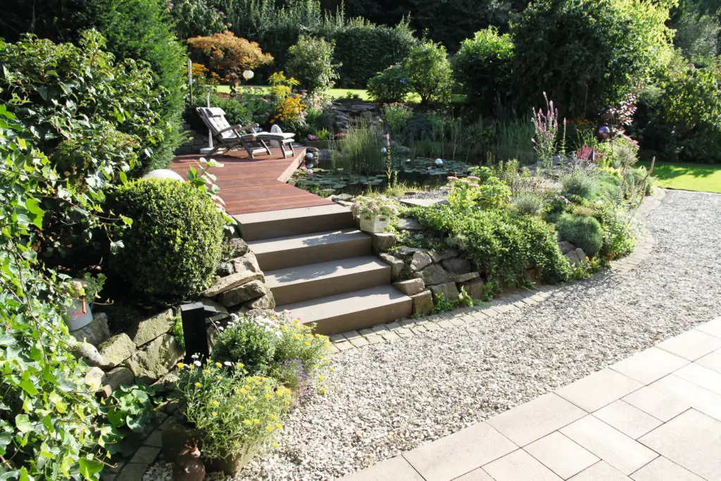 Ein angelegter Garten mit Steinstufen, die zu einer Holzterrasse mit zwei Liegestühlen führen, umgeben von Grünpflanzen, Blumen, Steinen und einem Kiesweg.