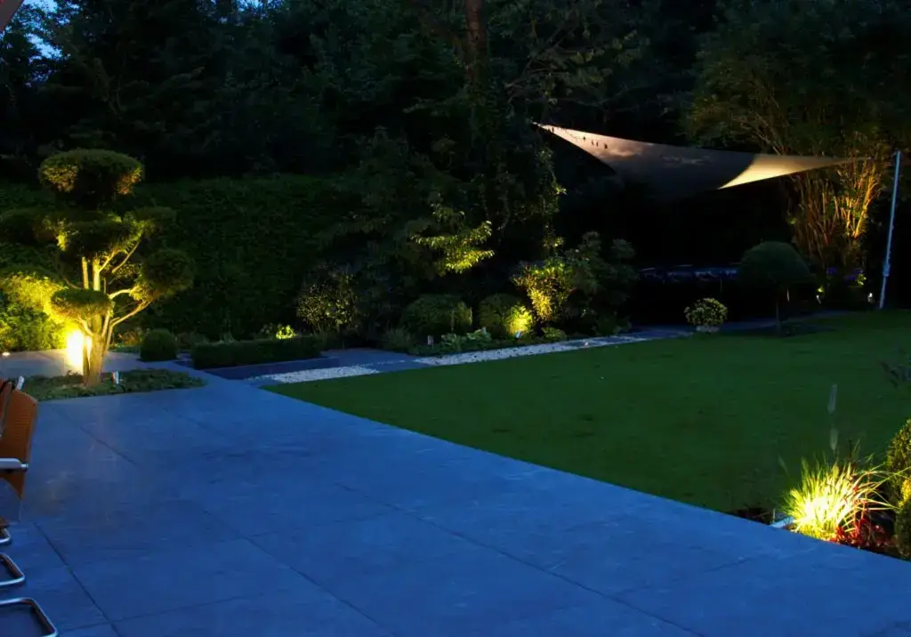 Ein moderner Garten in der Abenddämmerung mit einer großen gefliesten Terrasse, ordentlich geschnittenen Sträuchern und Bäumen, einer Rasenfläche und einer Gartenbeleuchtung, die die Pflanzen beleuchtet. Über einen Teil des Gartens ist ein Sonnensegel gespannt.