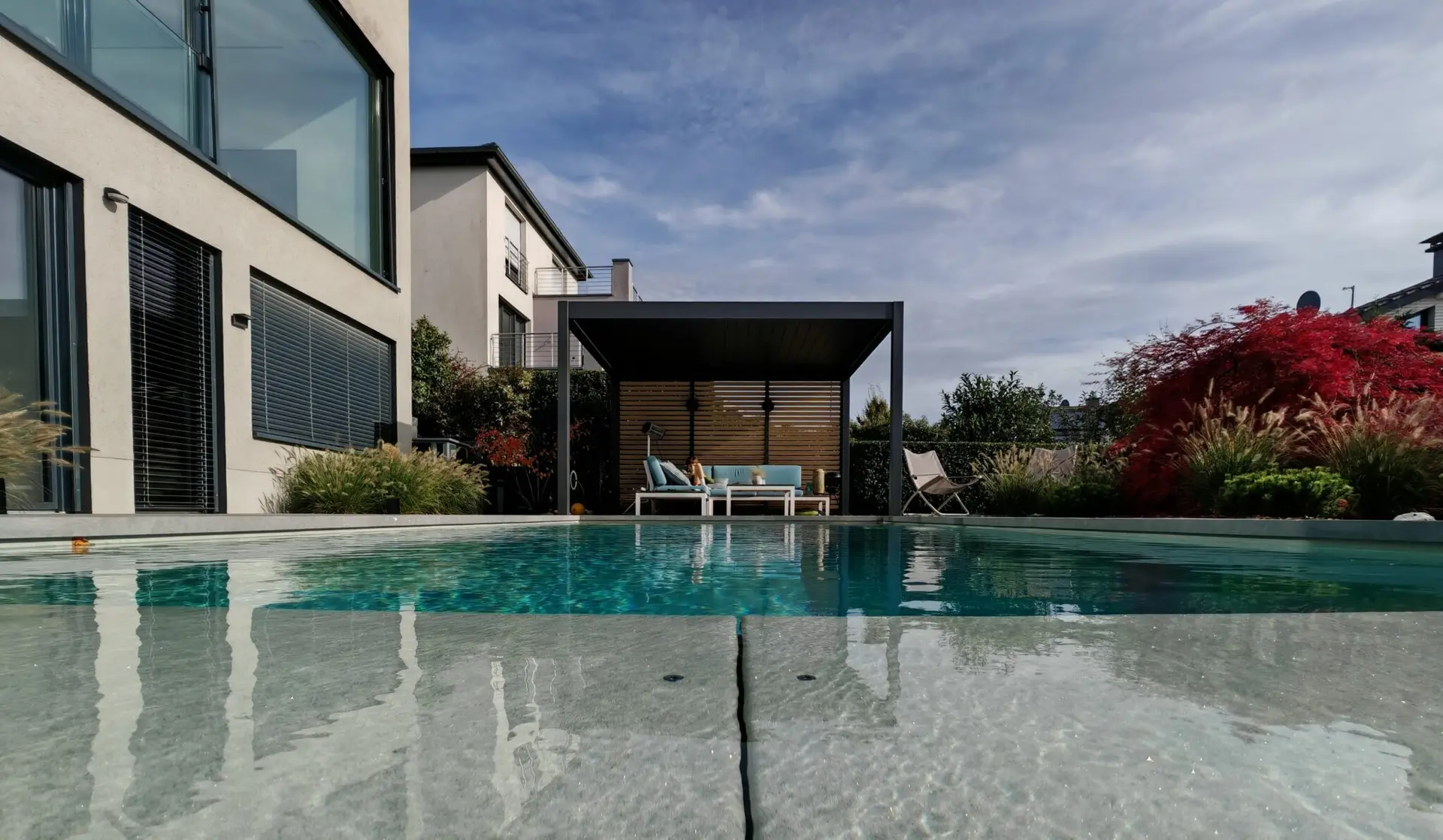 Ein modernes Haus mit großen Fenstern blickt auf einen Swimmingpool im Garten. Am anderen Ende des Pools gibt es einen schattigen Sitzbereich mit Gartenmöbeln. Dekorative Pflanzen und Sträucher umgeben den Platz.