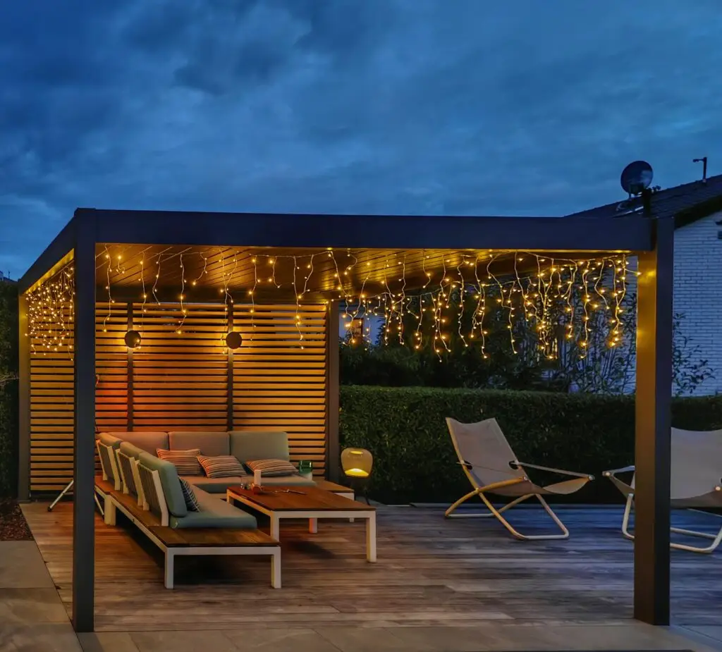 Eine moderne Außenterrasse in der Abenddämmerung mit einer Pergola mit Lichterketten, einem Sektionssofa mit Kissen, einem Couchtisch, Loungesesseln und Grünpflanzen im Hintergrund. Die Umgebung wird durch warmes gelbes Licht beleuchtet.