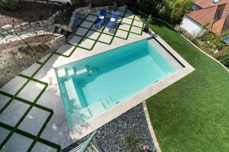 Ein rechteckiger Swimmingpool mit klarem Wasser, umgeben von Betonfliesen und grünen Grasstreifen, mit zwei Liegestühlen und einem kleinen Tisch an einer Seite und ordentlich angelegten Beeten.