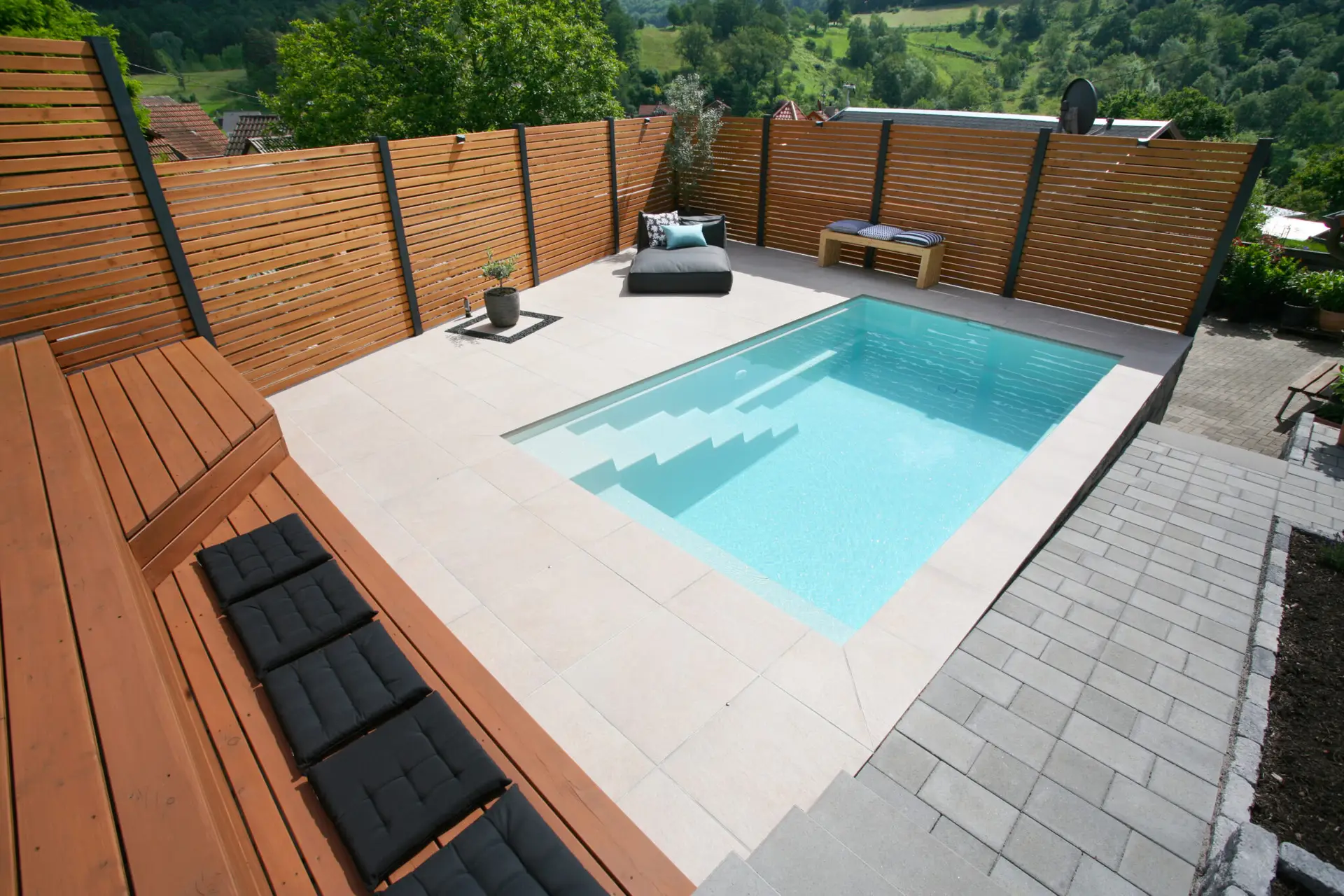 Ein kleiner rechteckiger Pool im Garten mit Stufen, umgeben von beigen Fliesen, einem Holzzaun und Holzbänken mit schwarzen Kissen. Eine einzelne Topfpflanze und ein Liegestuhl stehen in der Nähe des Pools. Hinter dem Zaun ist eine grüne Landschaft zu sehen.