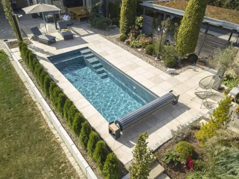 Ein rechteckiger, unterirdischer Swimmingpool mit klarem Wasser ist von beigen Fliesen, Liegestühlen, einem Sonnenschirm, Grünpflanzen und Gartenbeeten in einem modernen Hinterhof umgeben.