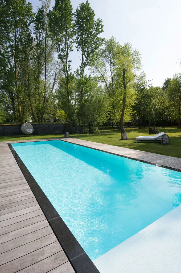 Ein rechteckiger Swimmingpool mit klarem, blauem Wasser ist von einem Holzdeck umgeben. Liegestühle und Bäume sind im grasbewachsenen Hof hinter dem Pool zu sehen. Die Szene ist hell und sonnig