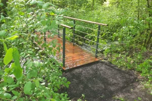 Ein kleiner Holzsteg mit Metallgeländer führt über einen schmalen Bach in einem üppigen, grünen Waldgebiet. Der Boden in der Nähe der Brücke scheint nass und schlammig zu sein.